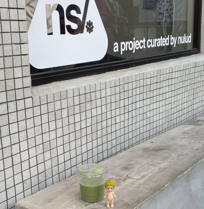 matcha nigiri 23 A minimalist outdoor setting featuring a matcha latte in a plastic cup alongside a small Sonny Angel figurine with a pineapple-themed headpiece. The scene is set against a tiled wall with a sign reading "ns/ a project curated by nulud."