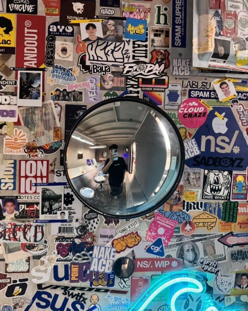 ns Sticker-covered wall at Nigiri Station Pampanga, featuring a convex mirror reflection of a person taking a hallway mirror selfie in an urban street-style setting.