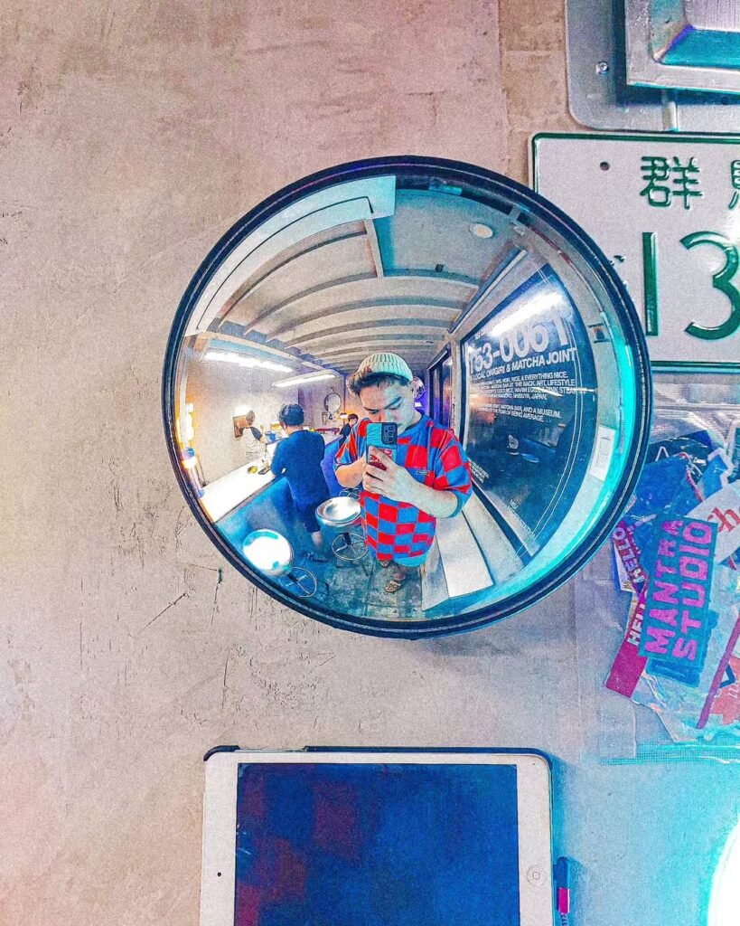 nss Circular convex mirror at Nigiri Station Pampanga, reflecting a person in a checkered outfit, set against a textured beige wall with neon lights in an urban setting.