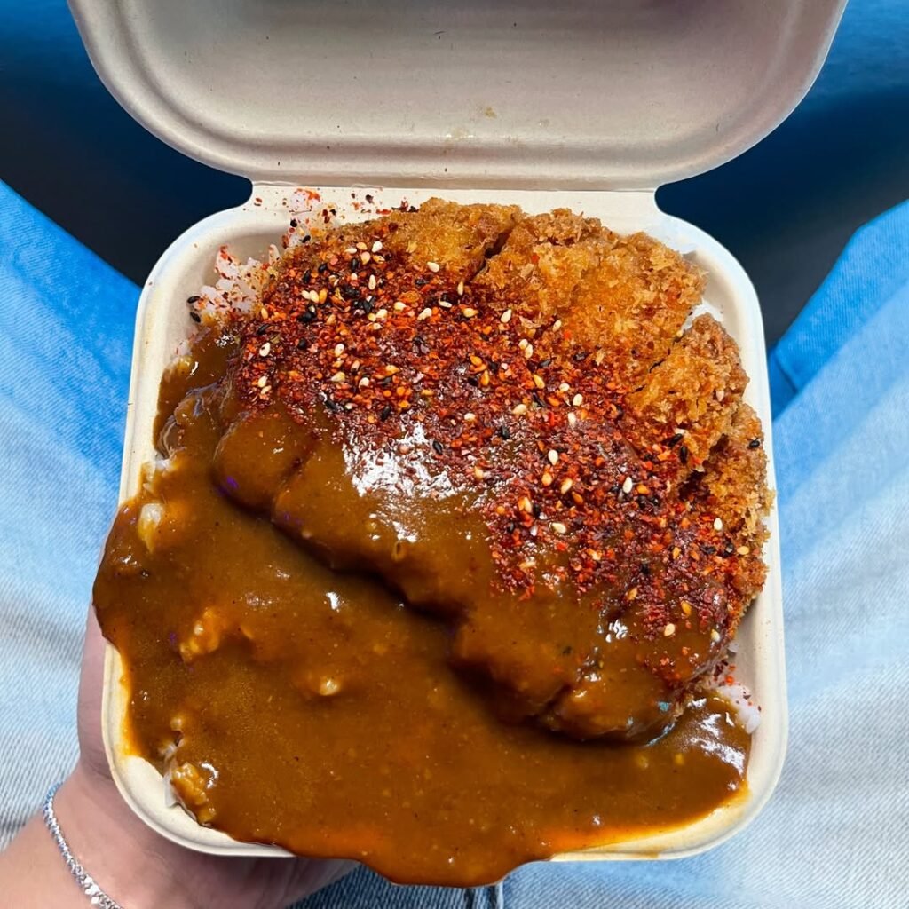 rice A crispy, golden-brown katsu cutlet, smothered in rich Japanese curry, topped with a bold chili spice mix, and served in a takeout container.